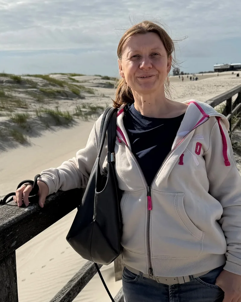 Kamila Diedrichs-Gern lehnt l&auml;chelnd am Holzgel&auml;nder eines Bohlenwegs zwischen D&uuml;nen am Meer